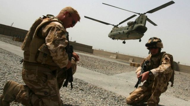 A Chinook helicopter can be seen taking off in the background with troops in the foreground